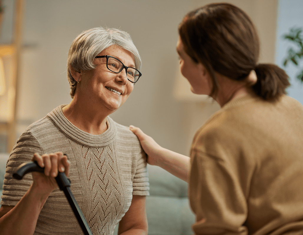 An elderly woman with gray hair and glasses smiling warmly as a caregiver or visitor gently touches her shoulder. The elderly woman holds a cane in one hand. The setting appears to be a care center or assisted living facility.