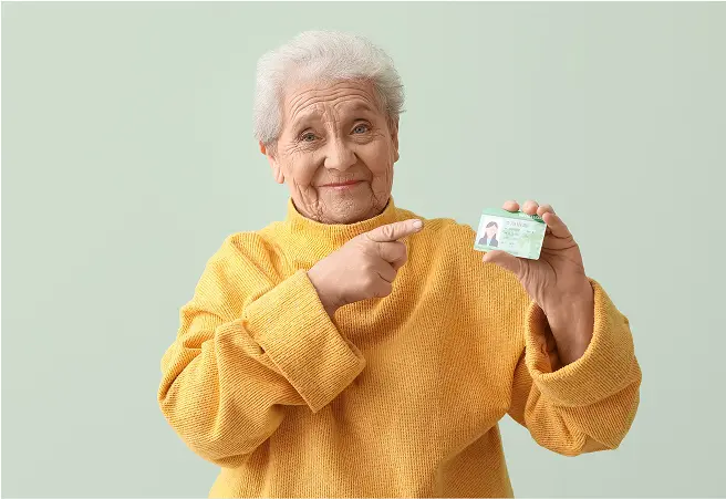 An elderly woman with gray hair, wearing a yellow sweater, smiling and pointing to a driver’s license she is holding in her right hand against a light green background.