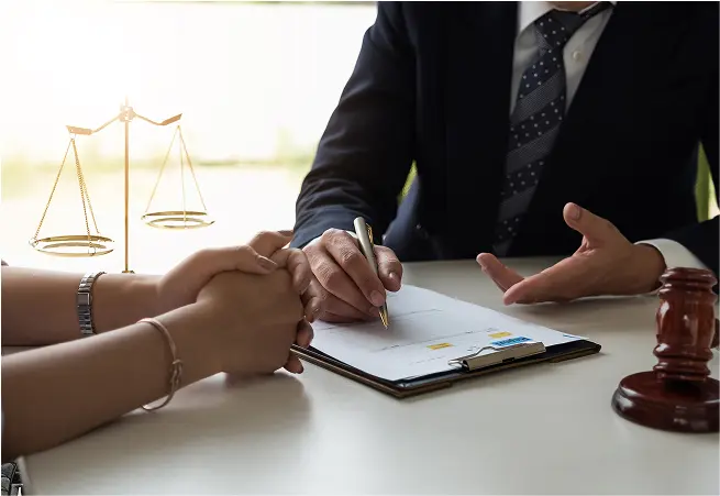 A person in a suit is signing a document at a table while another person holds their hands, with a gavel and a set of scales in the background, symbolizing legal or financial planning.