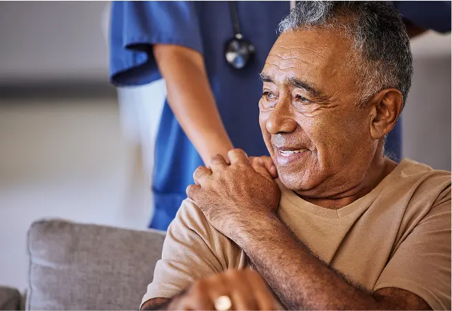 A smiling elderly man sitting on a couch, with a healthcare worker standing behind him. The man is wearing a beige shirt, and the healthcare worker is dressed in blue scrubs with a stethoscope. The man appears to be in a relaxed setting, possibly in a healthcare or care facility environment.