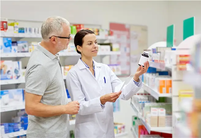 A female pharmacist showing a medication bottle to an older male customer in a pharmacy aisle filled with various medicine boxes and products.